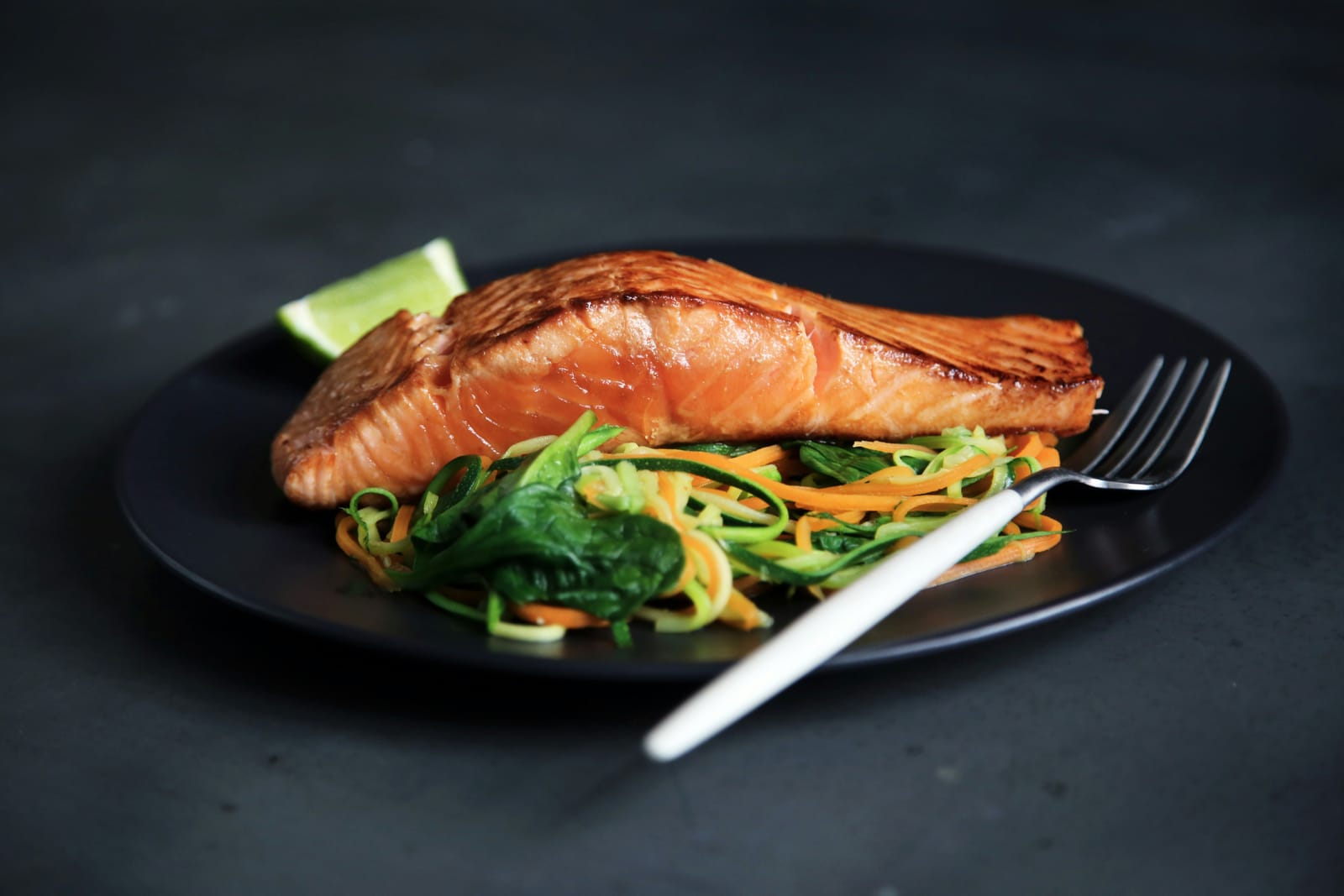 A cooked salmon fillet on a dark plate with courgette and carrot ribbons and a wedge of lime
