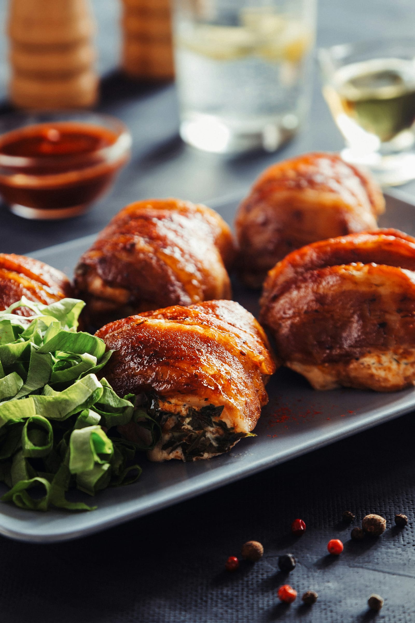 Bacon-wrapped stuffed chicken thighs on a dark plate with shredded greens and a small bowl of sauce