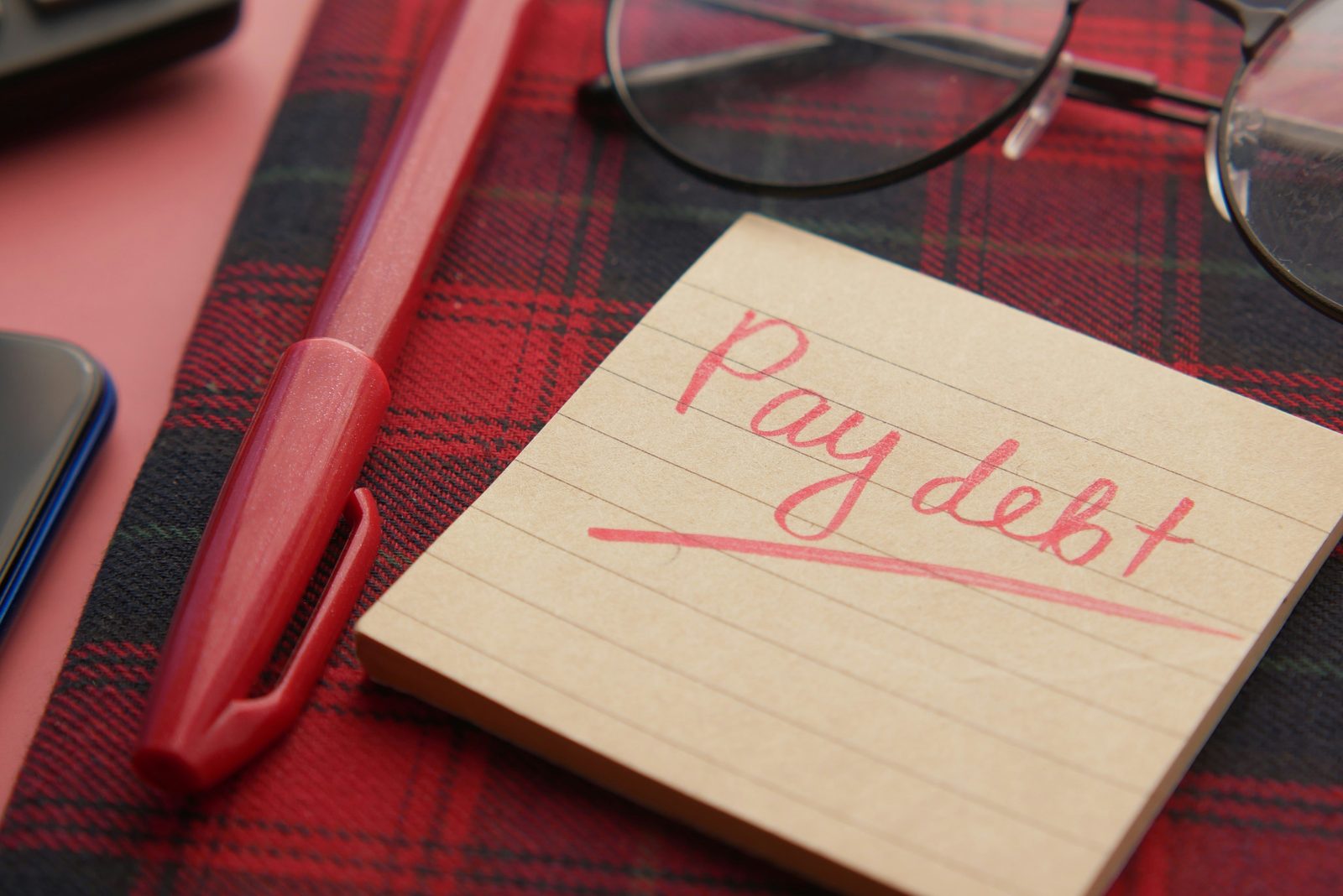 A notepad with the words Pay debt written and underlined in red, next to a pen and reading glasses
