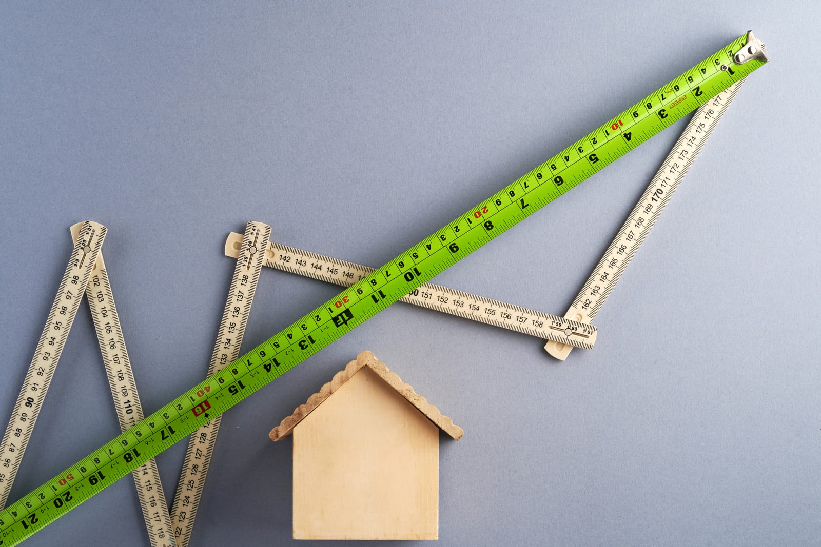 Folding wooden rulers and a green tape measure arranged in a rising zigzag above a small wooden model house on a grey background